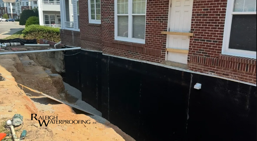 Professional foundation waterproofing installation on a Raleigh NC home showing black membrane applied to excavated foundation wall with red clay soil
